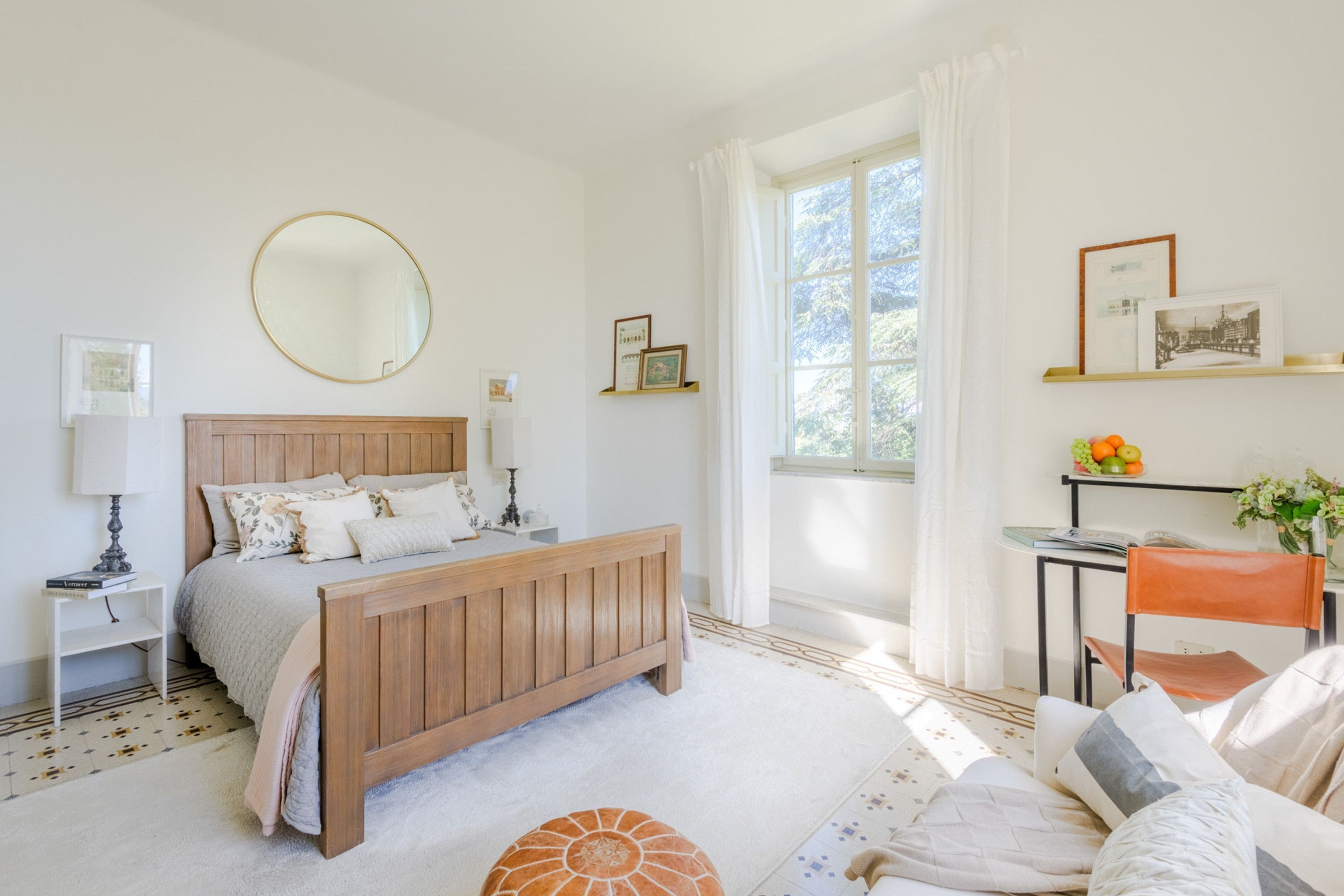 Un dormitorio con cama de madera, un espejo redondo, una mesita de noche con lámpara, una silla y una ventana con cortinas.
