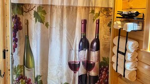 A room with a wooden ceiling, a curtain with grapevine and wine bottle prints, two wine bottles, two wine glasses, and a towel rack with towels.