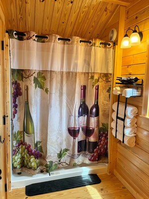 A room with a wooden ceiling, a curtain with grapevine and wine bottle prints, two wine bottles, two wine glasses, and a towel rack with towels.