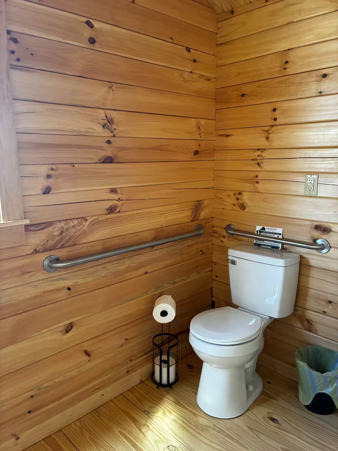 Un bagno con rivestimento in legno, un water bianco, un corrimano e un cestino.