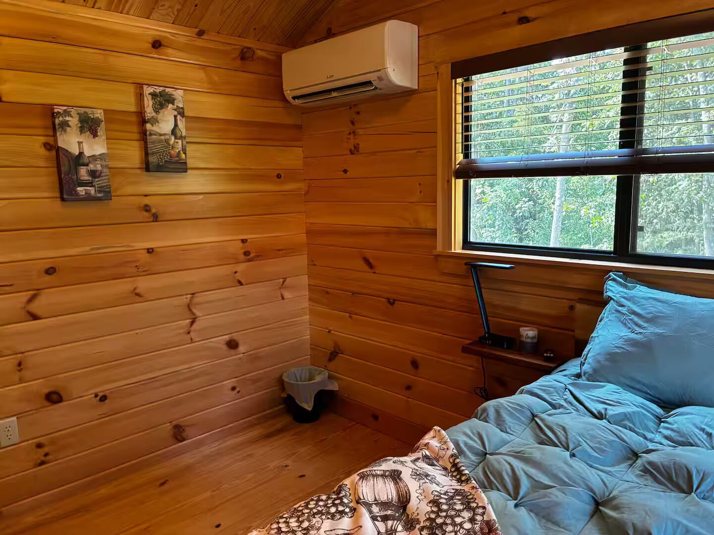 Ein Zimmer in einer Holzhütte mit einem Bett, einem Fenster mit Jalousien, einer Klimaanlage und zwei Bildern an der Wand.