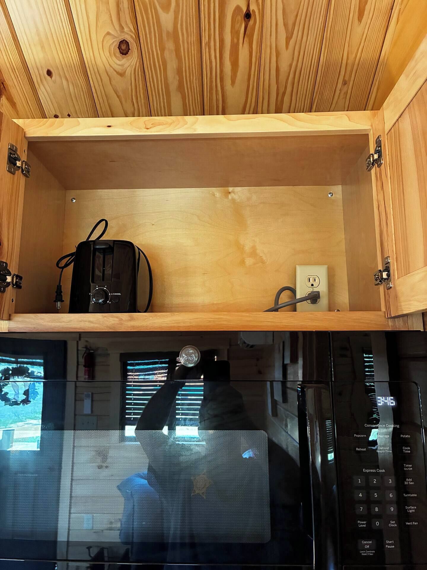 Een magnetron met een digitaal display en bedieningspaneel, een stopcontact en een klein plankje met een zwart apparaat en een stroomkabel.