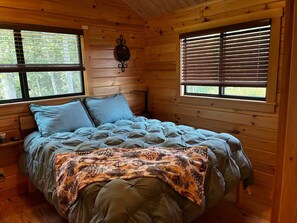 A wooden cabin bedroom with a bed, two windows with blinds, a wall-mounted light fixture, and a small nightstand.