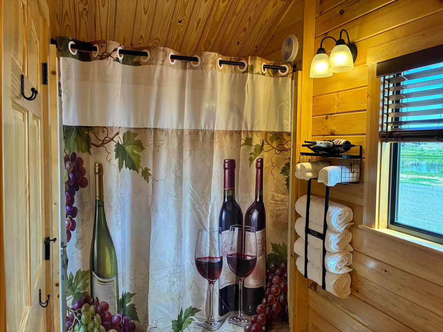 Ein Zimmer mit Holzvertäfelung, einem Fenster mit Jalousien, einem Vorhang mit Rebmuster und einem Regal mit Flaschen und Gläsern.