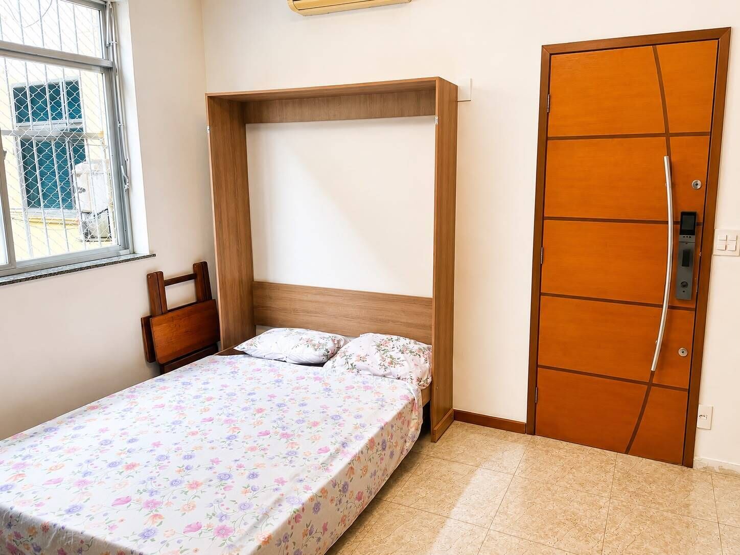 Quarto pequeno com cama de solteiro, cabeceira de madeira, janela com vista e porta com fechadura.