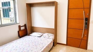 Quarto pequeno com cama de solteiro, cabeceira de madeira, janela com vista e porta com fechadura.