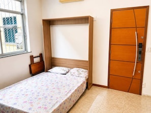 Una habitación pequeña con cama individual, cabecera de madera, ventana con vistas y puerta con cerradura.
