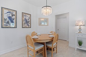 A dining area with a round table, four chairs, and a chandelier. There are three framed artworks on the wall. - 🌞🌴Pearl Street Retreat | Heated Pool + Steps to Beach (Fort Myers Beach)