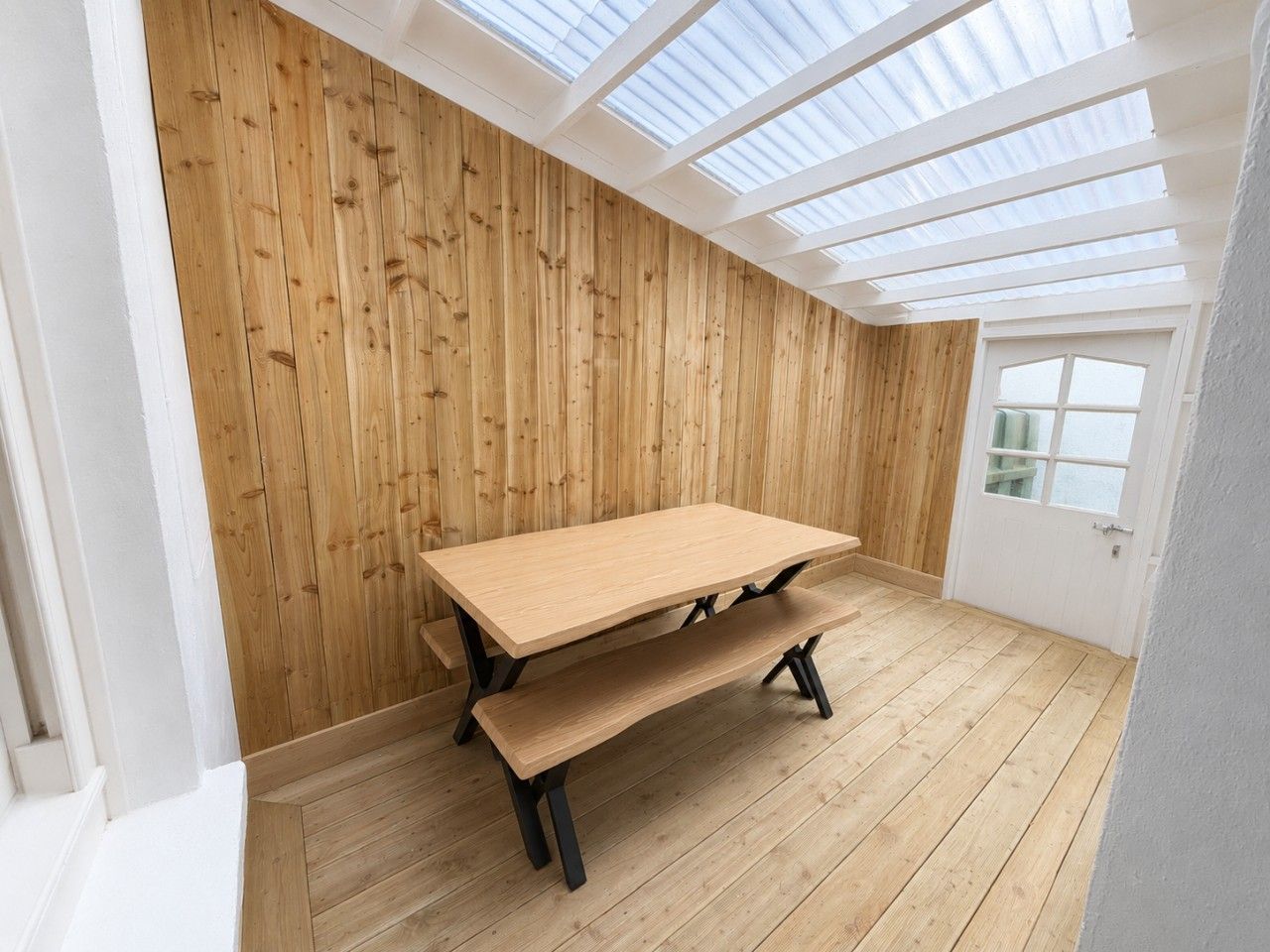 Une pièce avec une table et un banc en bois, une lucarne et une porte blanche.