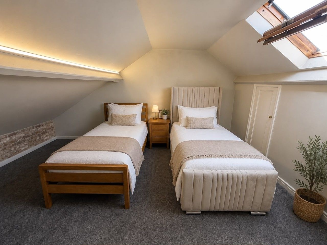 Ein Schlafzimmer mit zwei Betten, einem Oberlicht, einem kleinen Nachttisch mit Lampe und einem Blumentopf.