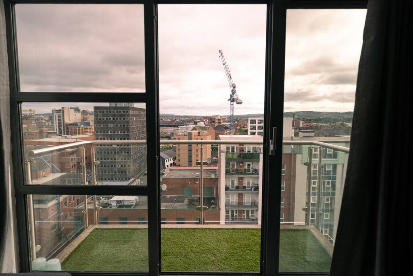 A view from a balcony with a grassy area, overlooking a cityscape with buildings and a construction crane.