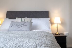Ein ordentlich bezogenes Bett mit einer flauschigen weißen Decke, einem Kissen mit dem Aufdruck 'snuggle' und einer Nachttischlampe auf einem Nachttisch.