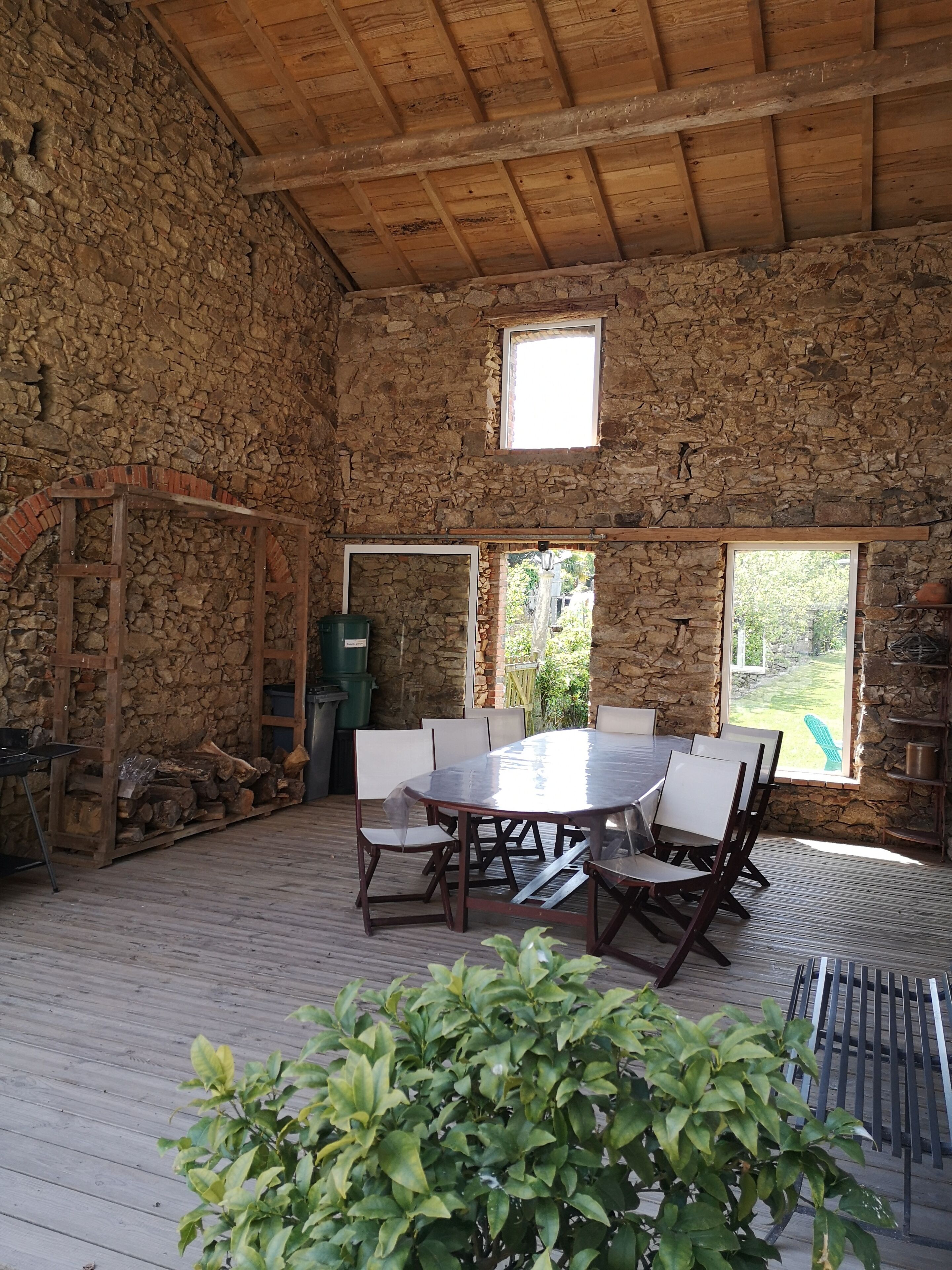 A rustic stone-walled room with a wooden ceiling, a large table with chairs, and two windows providing natural light.