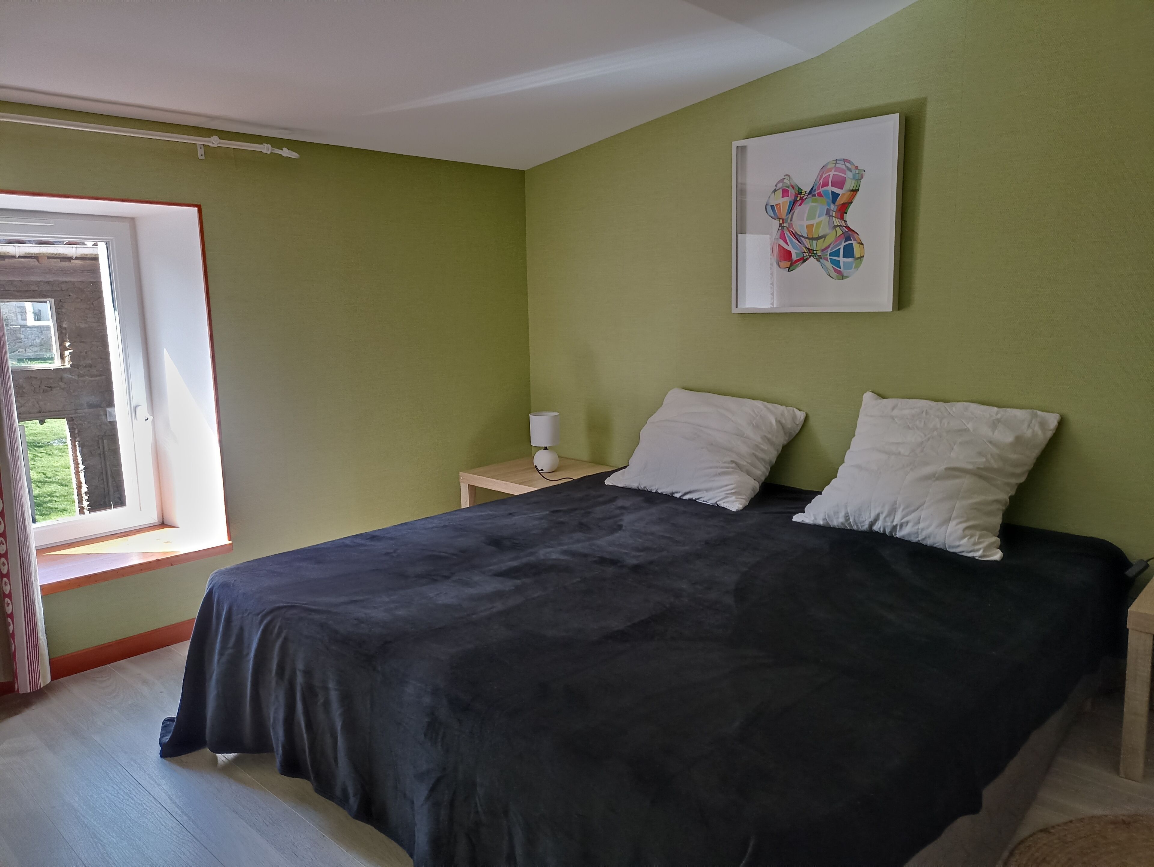 A bedroom with a bed, pillows, a bedside table with a lamp, a window, and a wall-mounted artwork.
