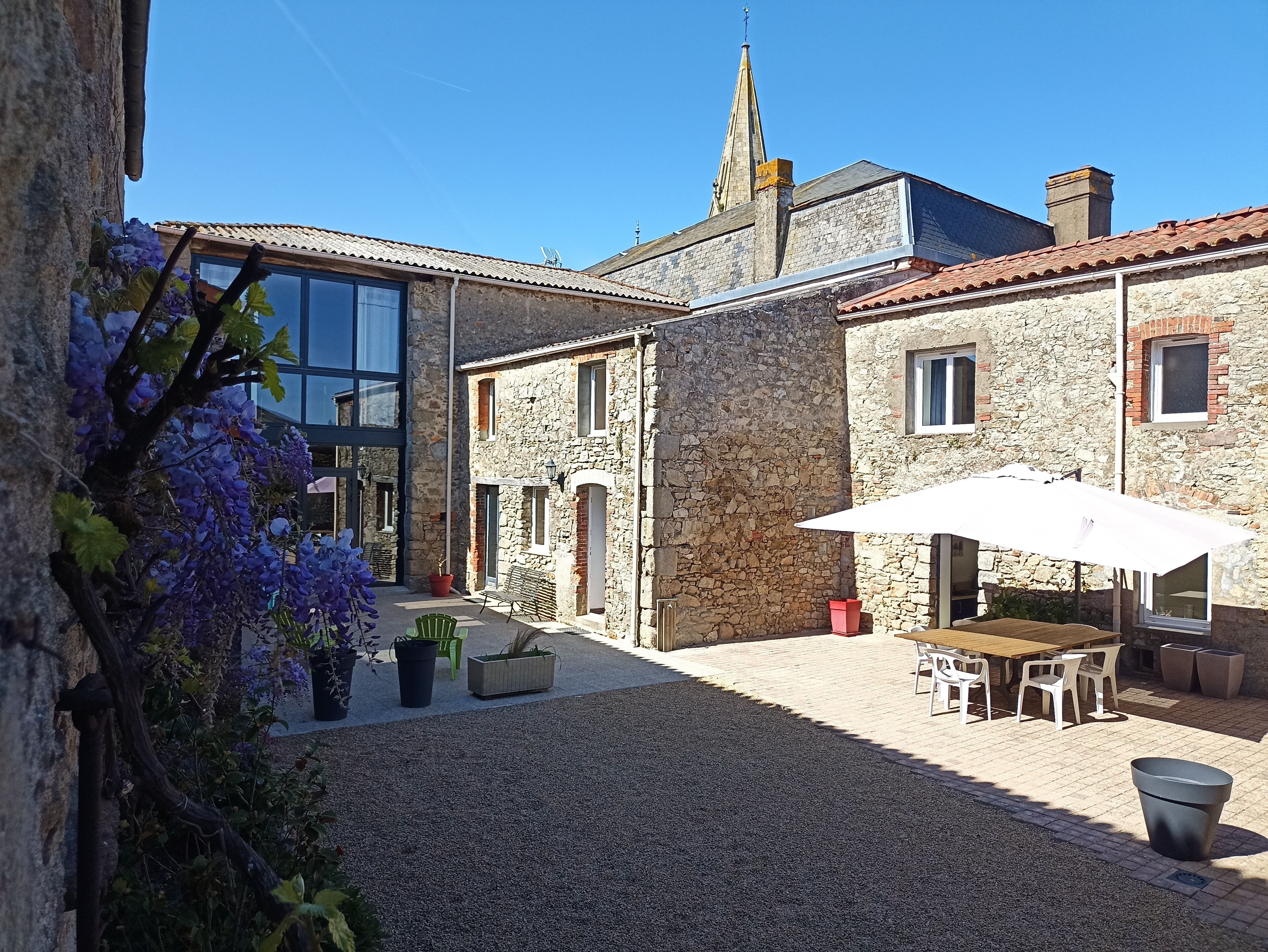 Une cour bordée de bâtiments en pierre, une pergola ornée de fleurs violettes, des sièges en plein air et, au loin, le clocher d’une église.