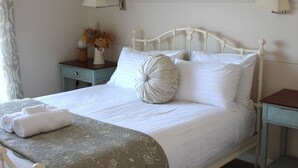 Een netjes opgemaakt bed met witte lakens en een decoratief kussen, een nachtkastje met een lamp en een vaas met gedroogde bloemen.