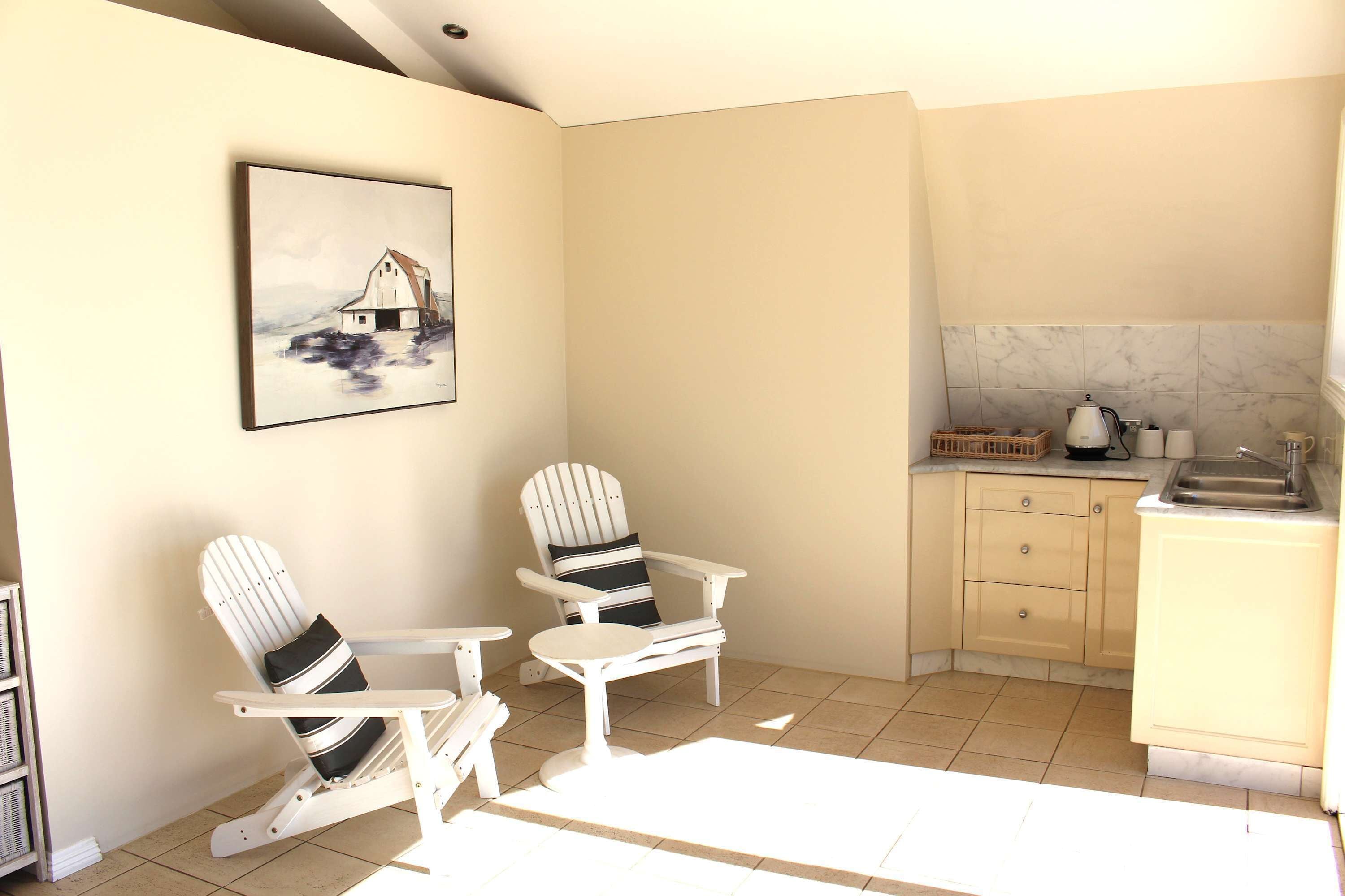 A room with two Adirondack chairs, a kitchen area with a sink and cabinets, and a painting of a barn on the wall.