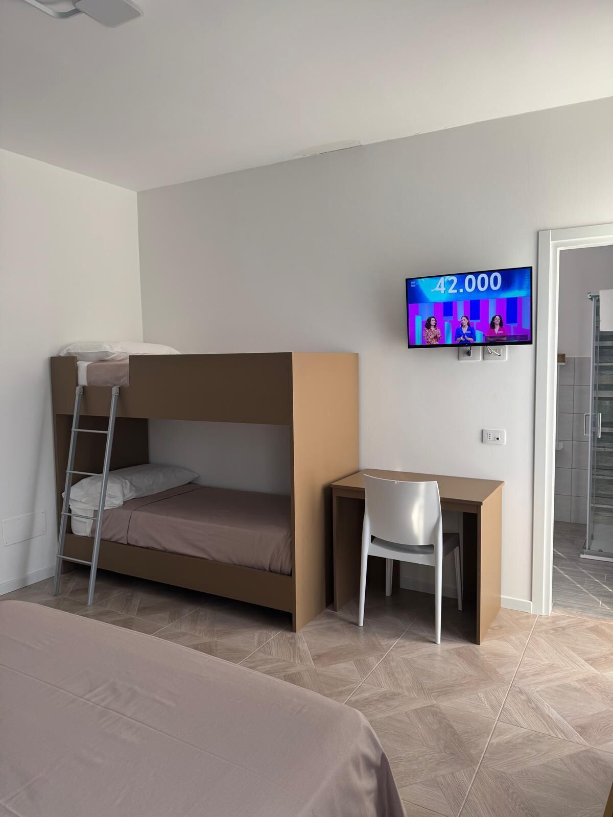 Una habitación con literas, un escritorio con silla, un televisor mostrando un programa de concursos y una zona de ducha.