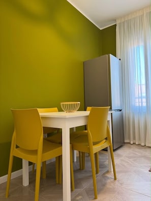 Un comedor con una mesa blanca y sillas amarillas, un bol blanco sobre la mesa, un refrigerador gris y una ventana con cortinas blancas.