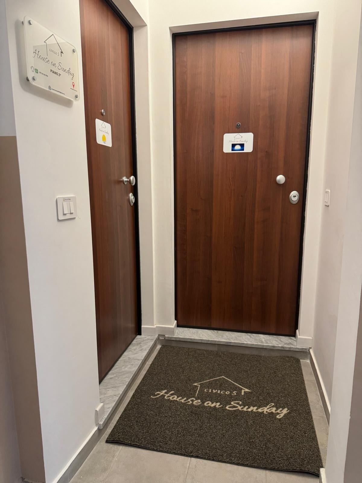 Um corredor com duas portas de madeira, um tapete de entrada com o texto 'House on Sunday' e uma placa indicando 'Civicos' e 'Piano 2'.