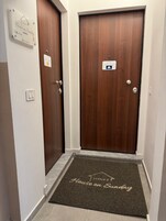 Un pasillo con dos puertas de madera, un felpudo con la inscripción 'House on Sunday' y un cartel que indica 'Civicos' y 'Piano 2'.