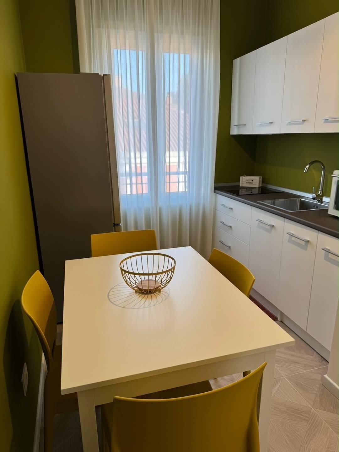 Eine kleine Küche mit Esstisch, Stühlen, einem Kühlschrank und einem Fenster mit Vorhängen.