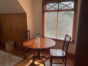 A dining area with a round table, chairs, a vase with flowers, a wooden cabinet, and a window with blinds.