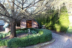 Un sendero de piedra conduce a un edificio rústico con puerta de madera, rodeado de exuberante vegetación y un jardín cuidado.