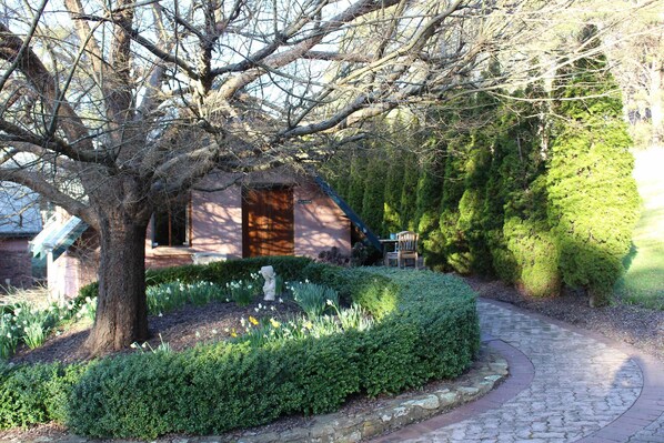 A stone pathway leading to a rustic building with a wooden door, surrounded by lush greenery and a well-maintained garden.