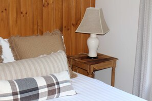 A bed with a white sheet, a plaid pillow, and two textured pillows. A table lamp is on a wooden nightstand beside the bed.