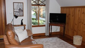 A living room with a leather sofa, a television on a wooden stand, a framed picture on the wall, and a window with a view of greenery. - Villa Marche - Taste of Tuscany (Riverside)