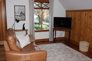A living room with a leather sofa, a television on a wooden stand, a framed picture on the wall, and a window with a view of greenery.