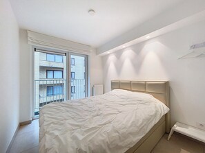 Ein modernes Schlafzimmer mit einem großen Bett, einem Fenster mit Blick auf ein Gebäude und einem weißen Nachttisch.