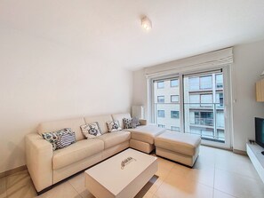 Ein modernes Wohnzimmer mit einem beigefarbenen Ecksofa, einem Couchtisch und einem großen Fenster mit Blick auf ein Gebäude.