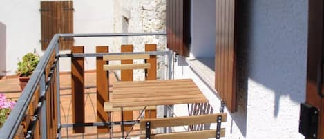 A balcony with a wooden table and chairs, a white wall with a window, and a door with a wooden shutter.