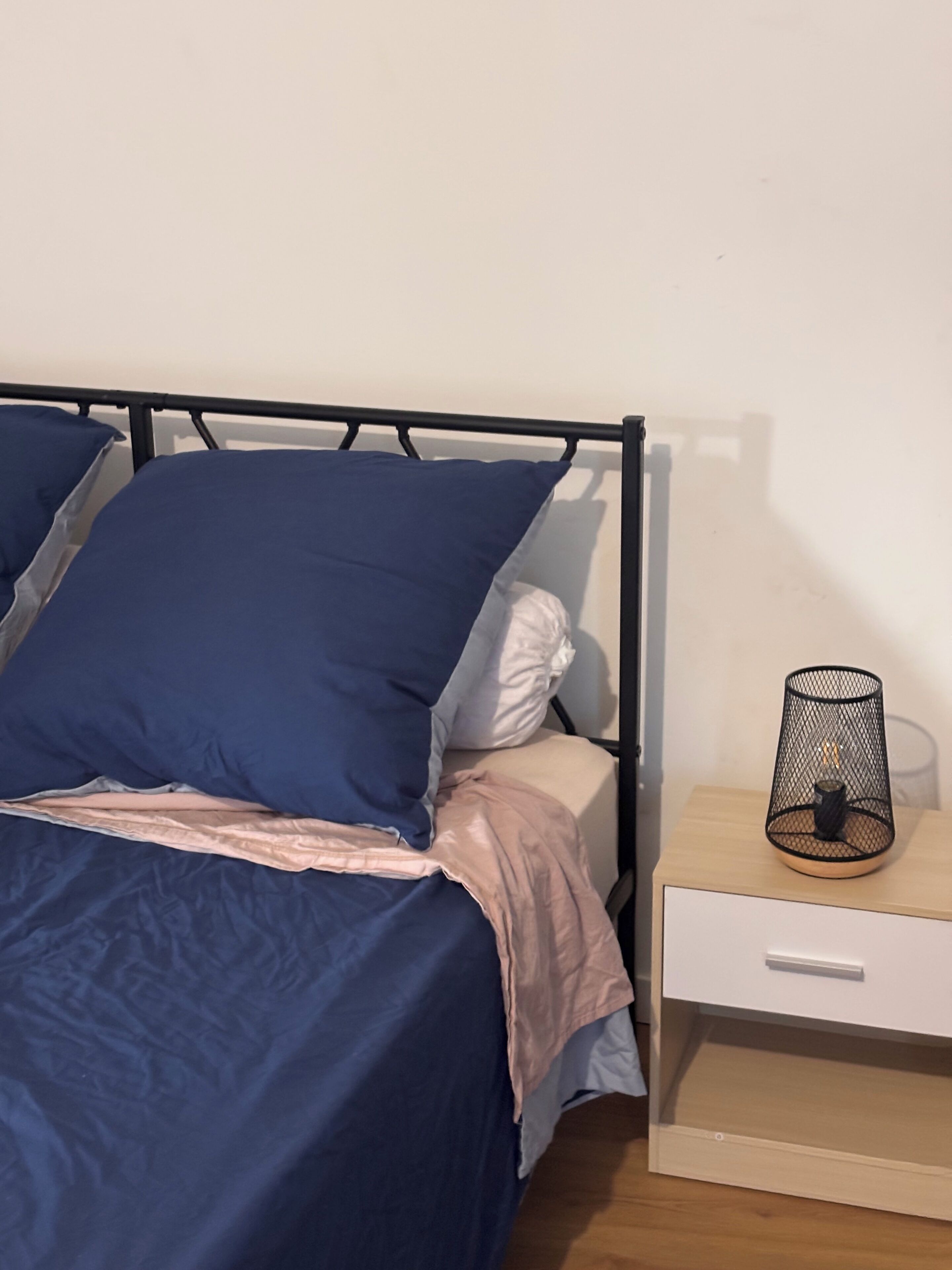 Een bed met een metalen frame, blauw beddengoed en een nachtkastje met een lamp.