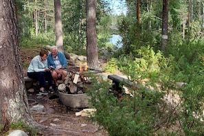 Two people sitting by a campfire in a forested area with a river in the background.