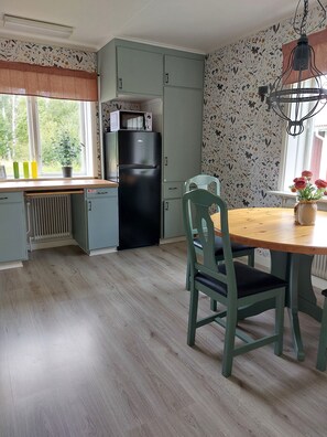 Een keuken met een koelkast, een magnetron, een tafel met stoelen en een raam met uitzicht op groen.