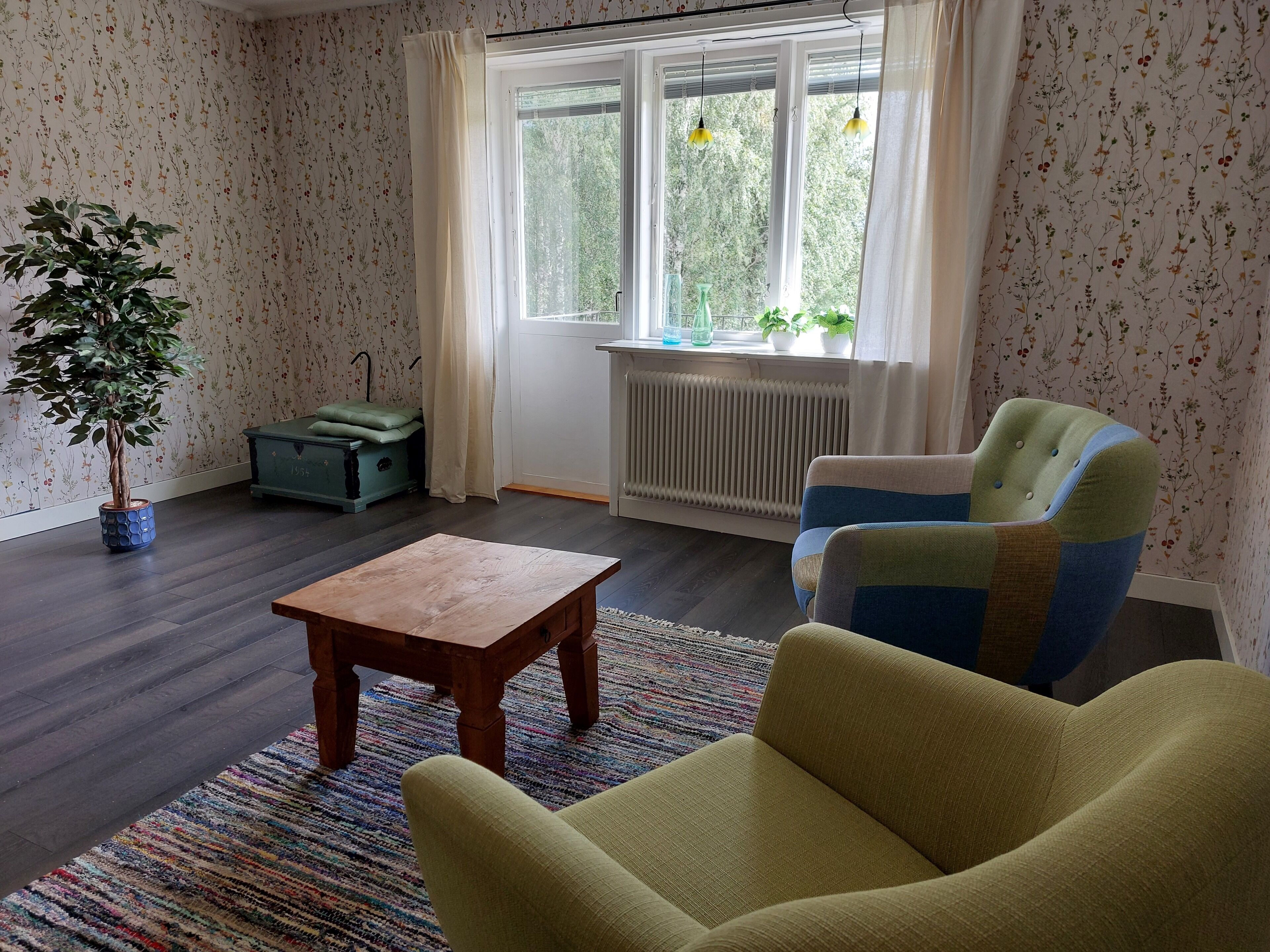 Ein Zimmer mit Holzboden, einem grünen Sessel, einem kleinen Holztisch, einem Heizkörper und einem Fenster mit Vorhängen.