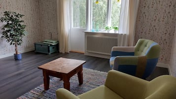 Ein Zimmer mit Holzboden, einem grünen Sessel, einem kleinen Holztisch, einem Heizkörper und einem Fenster mit Vorhängen.