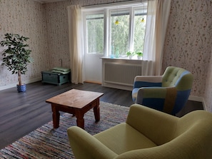 A room with a wooden floor, a green armchair, a small wooden table, a radiator, and a window with curtains.