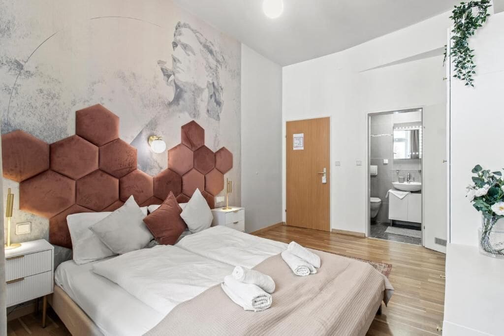 Habitación de hotel moderna con una cama grande, cabecera con diseño geométrico, mesita de noche con lámpara y vista al baño.