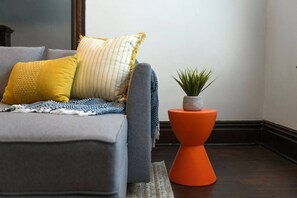 Ein graues Sofa mit gelben und gestreiften Kissen, einer blau-weißen Decke und einem kleinen orangefarbenen Beistelltisch mit einer Pflanze.