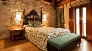 A bedroom with a stone wall, wooden ceiling beams, a bed with a patterned bedspread, a bench, and a nightstand.