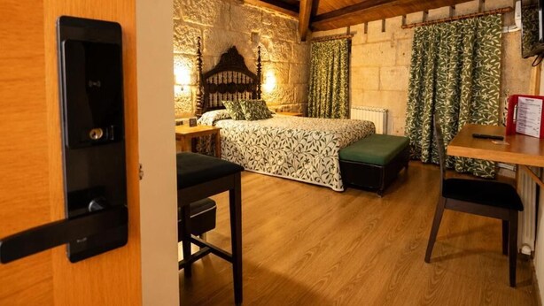 A room with a bed, a desk, and a chair. There is a wooden ceiling and a stone wall.