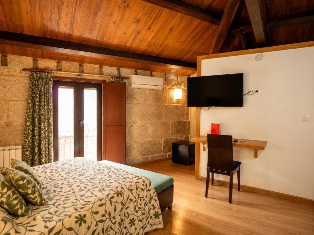 A bedroom with a bed, a television mounted on the wall, a desk with a chair, and a stone wall.