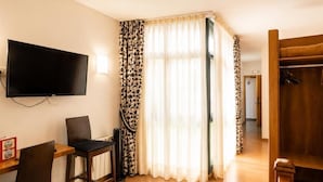 A room with a flat-screen TV mounted on the wall, a wooden desk with two chairs, a bed with a patterned bedspread, and a wardrobe with a mirror.
