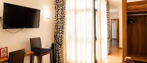 Habitación con televisión de pantalla plana montada en la pared, un escritorio de madera con dos sillas, una cama con colcha estampada y un armario con espejo.