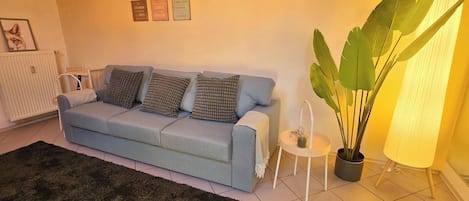Una sala de estar con un sofá azul claro, cojines estampados, una mesita auxiliar con una vela y una planta verde grande.
