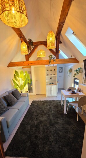 A cozy living room with a sofa, a coffee table, and a dining area with a table and chairs. There are wicker lamps hanging from the ceiling and a large plant in the corner. - Apartment Mutterstadt Oasis One - MIS (Mutterstadt)